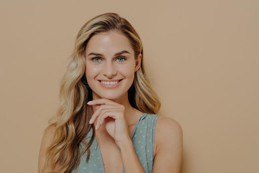 Pensive attractive young blonde female wearing blue dress holding hand under chin, smiling and looking at camera with happy face expression while standing isolated next to orange background