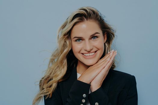 Portrait of young smiling joyful blondefemale student resting head on hands, expressing positive emotions with her gesture, experiencing happiness while posing isolated on blue background