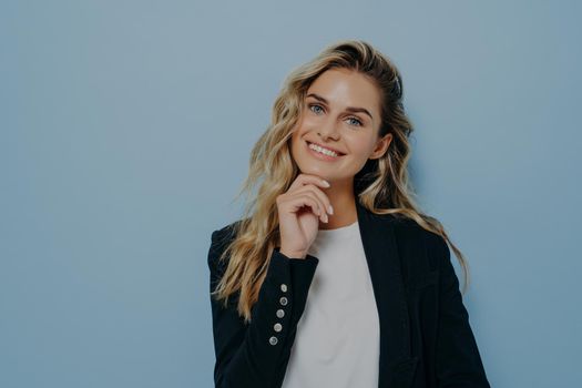 Young cute blonde woman touching with hand her chin, looking straight while smiling with big smile, making joyful and positive expression, posing isolated on blue background with copy space for text