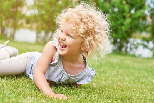 Shot of a cute little girl with blonde curly angelic hair lying on the grass laughing happily looking away copyspace nature enjoyment emotions kids children family recreation summer concept.