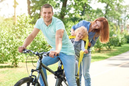 Happy young man smiling to the camera while cycling with his child in a baby bike seat and his beautiful wife at the local park on a warm summer day family active lifestyle concept.