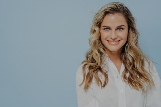 Attractive blonde woman in white shirt smiling and looking straight at camera with toothy smile and being calm at same time, posing alone next against blue background with copy space for text