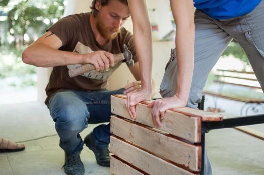 13.08.2021 - Ukraine, Goshcha, group of young workers is using power screwdriver drilling, hambler during construction wooden bench, do it together.