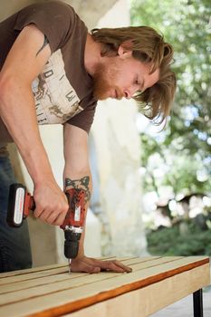 13.08.2021 - Ukraine, Goshcha, voluntary event, skilled young male worker is using power screwdriver drilling during construction wooden bench, do it yourself.