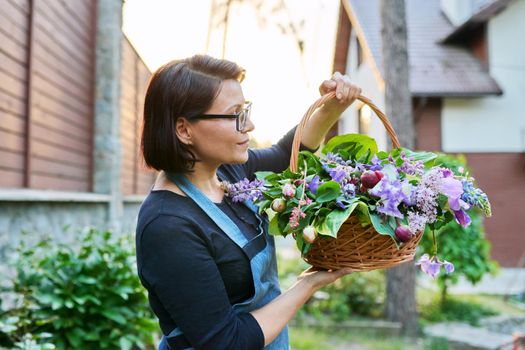 Middle-aged woman gardener florist in an apron with basket of fresh flowers folded in floral arrangement. Hobby, leisure, floristry, nature, beauty, gardening, creative bouquet, holiday concept