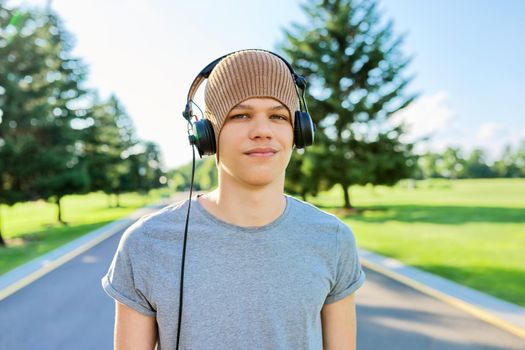Portrait of teenage hipster in knitted hat with headphones looking in camera. Smiling guy listening to music, summer green nature in park. Education, leisure, adolescence, lifestyle and technology