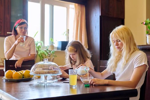 Family, girls children eating at table in kitchen. Home kitchen interior, mother with glass of milk, sisters eating and looking at smartphone screen. Lifestyle, daily routine, home, people, technology
