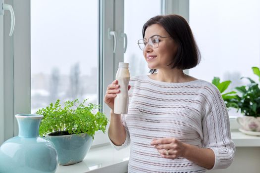 Middle-aged woman drinking milk drink, liquid yogurt in a bottle, at home near the window. Healthy food, lifestyle, winter season, mature age, nutrition, diet concept