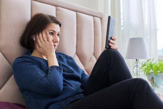 Sad middle aged woman in headphones looking into the smartphone screen. A frustrated female sitting at home in bed watching an unpleasant video. Technology, emotions, lifestyle, mature age concept