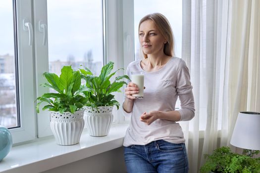Middle-aged woman with a glass of milk, at home near the window, in the autumn winter season. Health, beauty, lifestyle, food, mature age concept