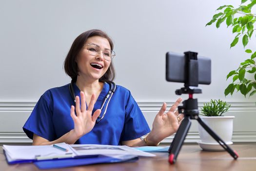 Female doctor with stethoscope consulting online using smartphone on tripod. Talking doctor looking at phone webcam, video meeting with patient. Medicine, technology, lifestyle, profession, people concept