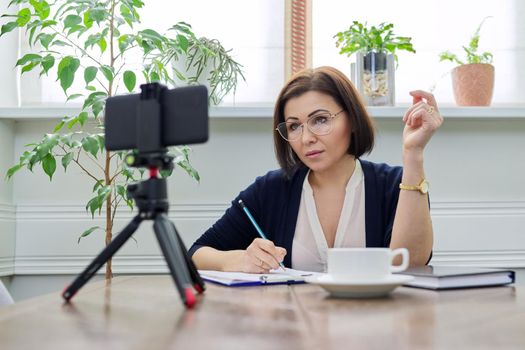 Middle-aged woman teacher, business woman working online using smartphone. Business confident female with papers on table talking looking in webcam phone on tripod, at home. Technology education work