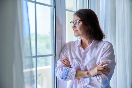 Portrait of a mature confident woman looking out the window. Middle-aged female wearing glasses with crossed arms, calm, smiling, resting