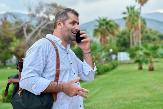 Mature business man talking on the phone outdoors. Serious confident male with backpack, in tropical park. Business trip, freelance, communication, technology, middle aged people concept, copy space
