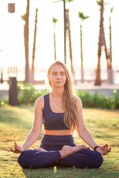 Young girl sitting in Lotus pose in the park near seafront line at sunset sunrise. Beautiful girl practicing Yoga during summer Vacation Meditation Seaside Sea Ocean Holiday Travel