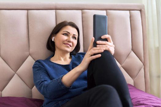 Smiling middle-aged woman in headphones looking in smartphone, resting female lying in bed at home, video on the phone, film, vlog, social network. Lifestyle, rest, technology, mature people concept