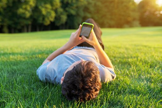 Guy teenager lying on grass with smartphone. Male in gray T-shirt with phone in his hands, green lawn background. Youth, technology, lifestyle, adolescence concept