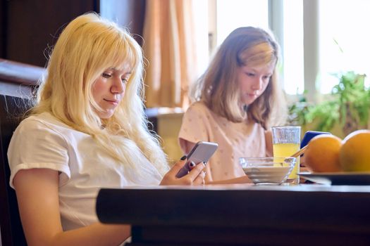 Children two girls sisters eating at home, looking at smartphones. Teenagers having breakfast in kitchen, using phones for leisure entertainment education. Technology in life lifestyle, adolescence