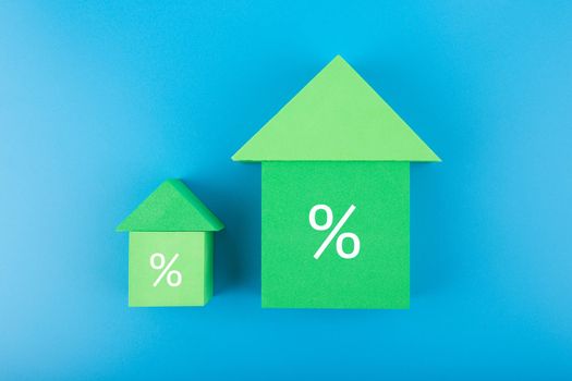 Two houses made of green toy blocks with increasing percentage sign on blue background. Modern concept of inflation, percent, mortgage or interest rate growth