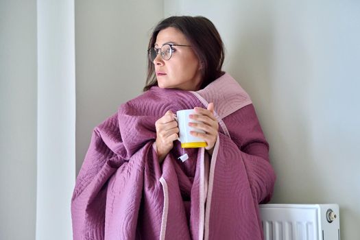 Cold winter autumn season at home, frozen woman basking herself with a warm blanket and a mug of tea near the central heating radiator