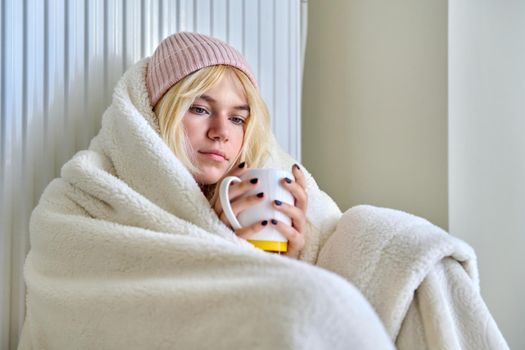 Heating during the cold winter autumn season. Young female teenager woman warming herself with a blanket hat, a central heating radiator, hot tea