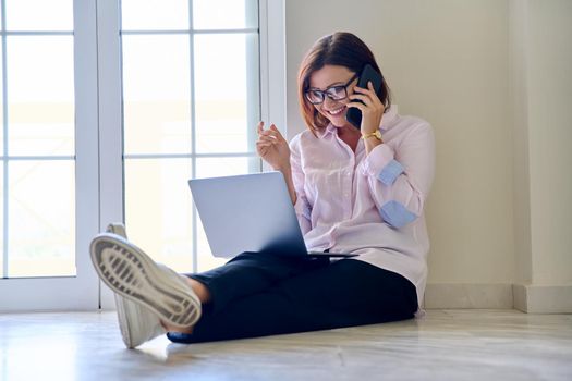 Business middle aged woman sitting on the floor with a laptop talking on a mobile phone. Business, technology, freelance, remote work, communication, home office, mature people concept