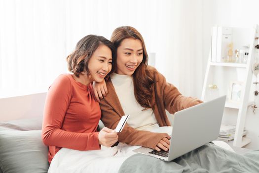 Two friends shopping online with credit card and laptop lying on the bed at home