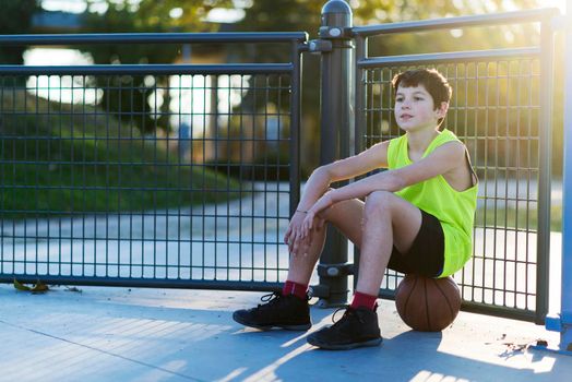 A young outdoors sitting on basket ball at a street court with ball
