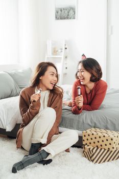 Cheerful young female friend with brush as mic singing while sitting on the bedwhile resting on bed during makeup