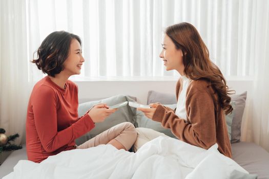 Smiling Two Asian young lovely couple repose on white bed and happy playing with smartphone and social media.