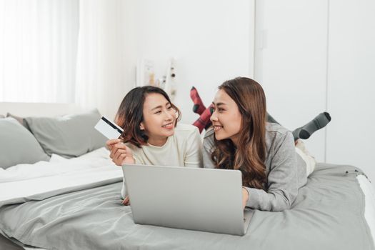 Two beautiful women lying in bed, holding a credit card and a laptop in it