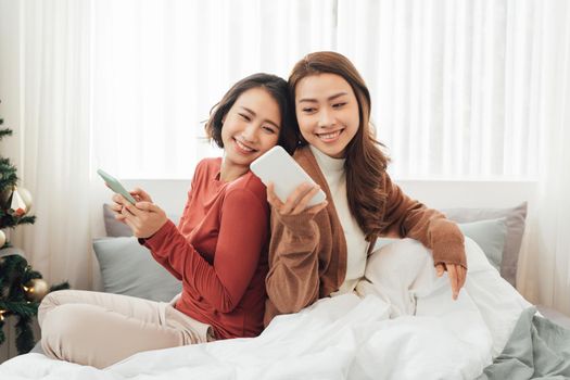 Smiling Two Asian young lovely couple repose on white bed and happy playing with smartphone and social media.