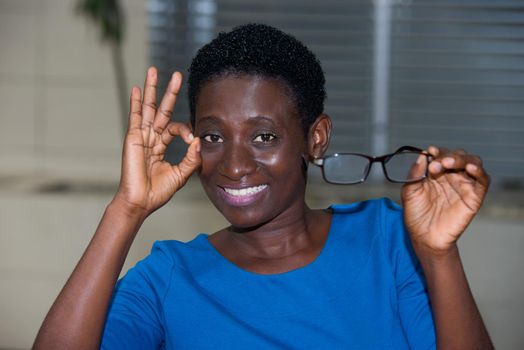 young girl standing in blue camisole eyeglasses in hand watching the camera smiling.