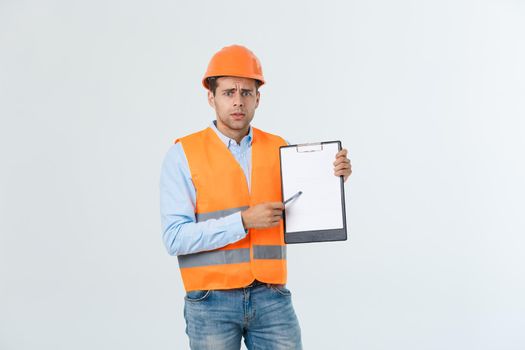 Frustrated young engineer with hardhat and reflective vest checking on mistake in document over gray background.