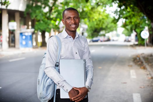 young man standing in street with laptop looking at the camera smiling.