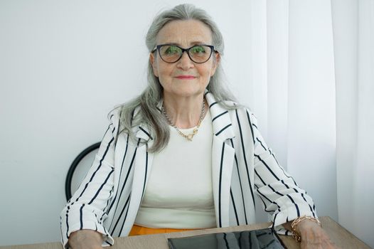 Senior grey haired businesswoman in striped jacket with eyeglasses is working in her office sitting at the desk. Happy retirement, employment and labor.