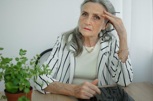 Tired senior grey haired businesswoman in striped jacket with eyeglasses is working in her office sitting at the desk and feeling bad due to menopause, menopause relief concept.