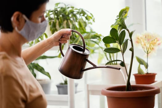 side view woman with face mask using watering can indoor plant. High resolution photo