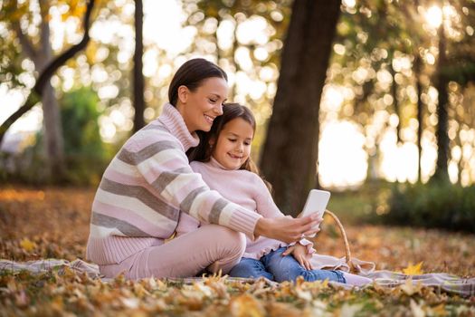 Mother and daughter using smartphone in autumn in park. They are taking selfie.