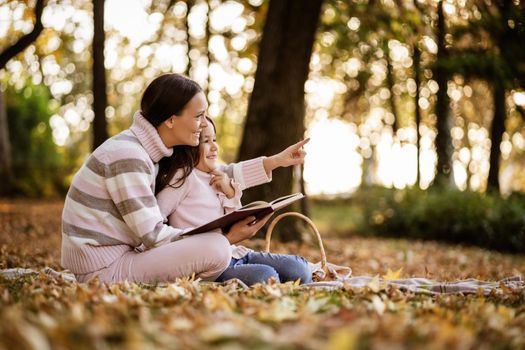 Mother and daughter enjoying autumn in park. Little girl is learning to read.