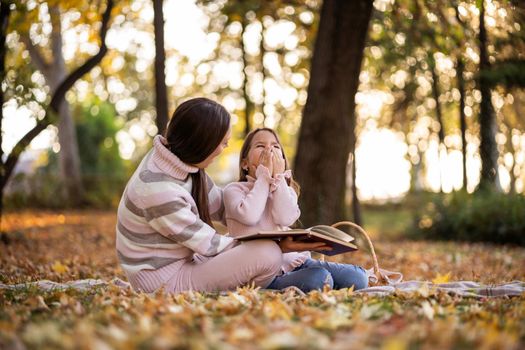 Mother and daughter enjoying autumn in park. Little girl is learning to read.