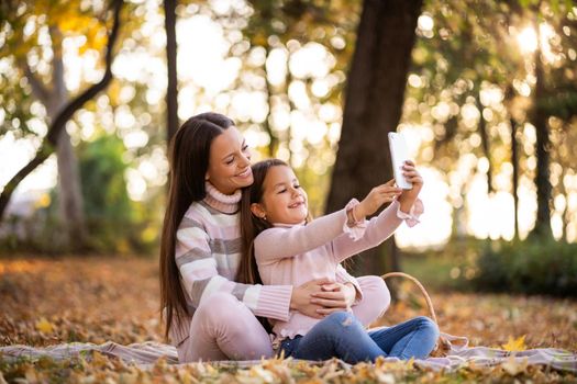 Mother and daughter using smartphone in autumn in park. They are taking selfie.