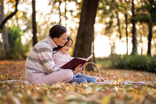 Mother and daughter enjoying autumn in park. Little girl is learning to read.