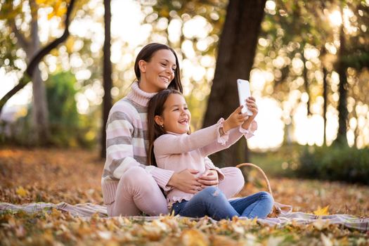 Mother and daughter using smartphone in autumn in park. They are taking selfie.