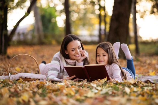 Mother and daughter enjoying autumn in park. Little girl is learning to read.