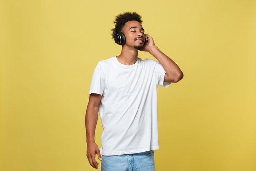 Close up portrait of dark skinned man with closed eyes, has headphones, listens to favourite music. Teenager enjoys audio track isolated on yellow background