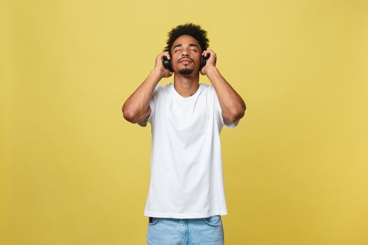 Close up portrait of dark skinned man with closed eyes, has headphones, listens to favourite music. Teenager enjoys audio track isolated on yellow background