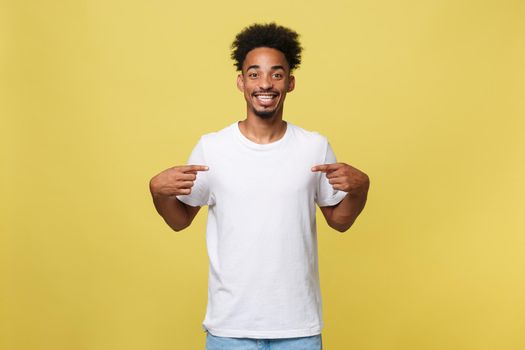 Portrait of handsome cheerful dark skinned male seller of clothes, dressed in casual white t shirt, indicates at blank copy space for your logo, isolated on yellow background. People, clothing, design.