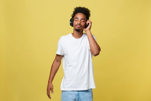 Close up portrait of dark skinned man with closed eyes, has headphones, listens to favourite music. Teenager enjoys audio track isolated on yellow background