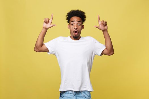 Astonished young African American man dressed in casual white shirt having excited fascinated look, pointing index finger at copy space on golden yellow background for your text or promotional content.